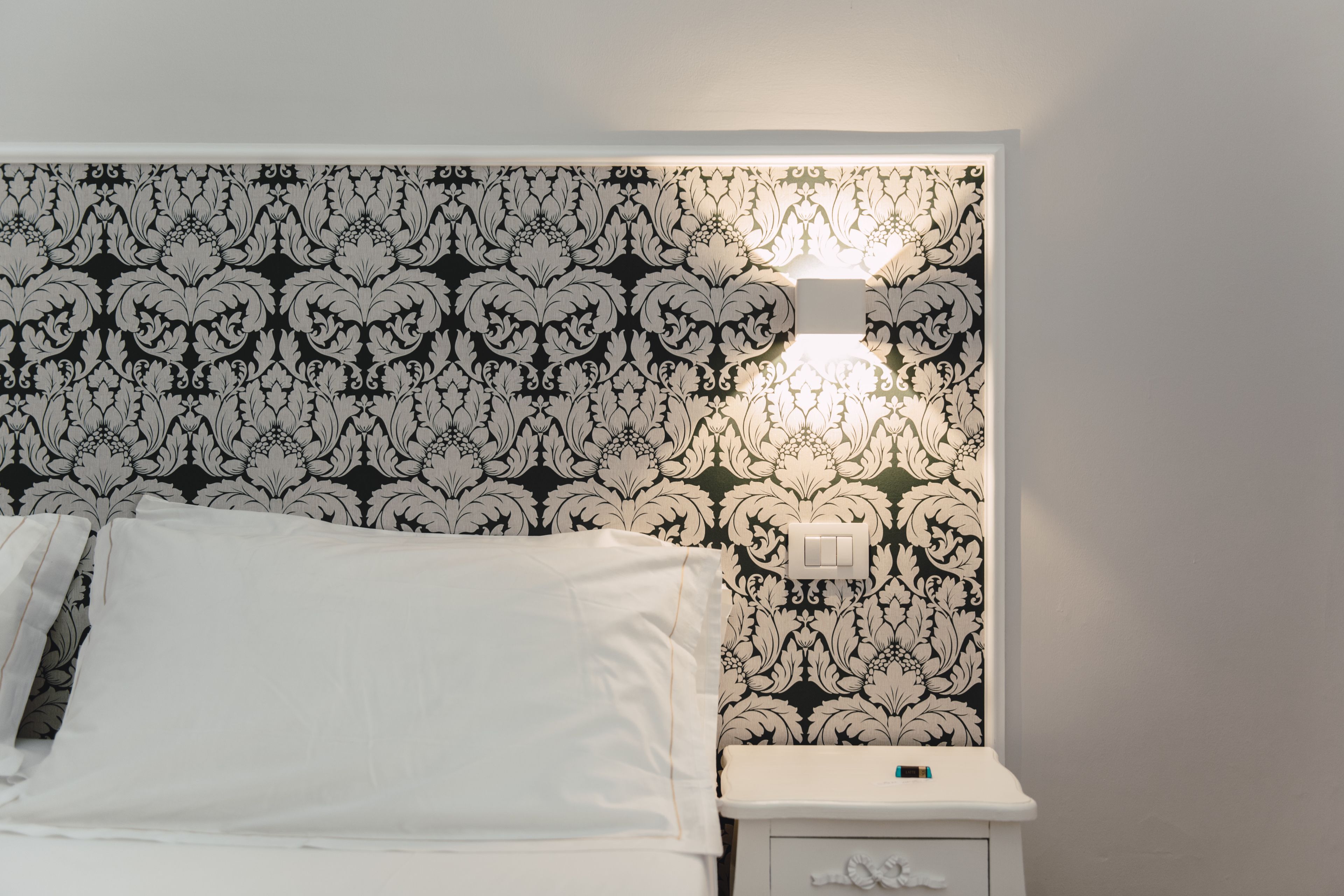 A neatly made bed with white linens, a bedside table with a lamp, and a wall with a black and white floral pattern.