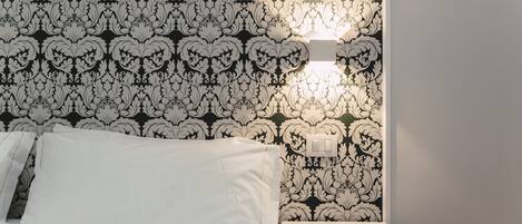 Una cama bien tendida con sábanas blancas, una mesita de noche con lámpara y una pared con estampado floral en blanco y negro.