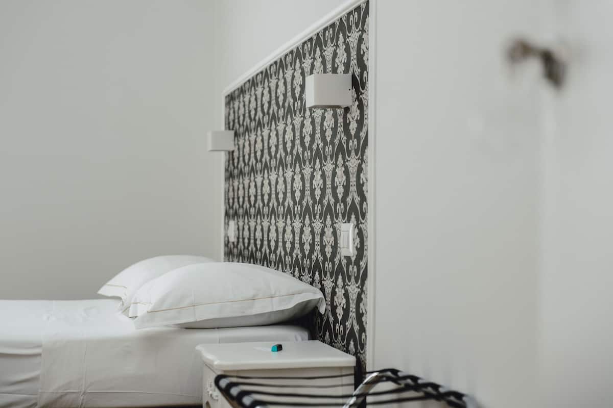 A neatly made bed with white linens and pillows, a bedside table with a lamp, and a patterned wallpaper on the wall.