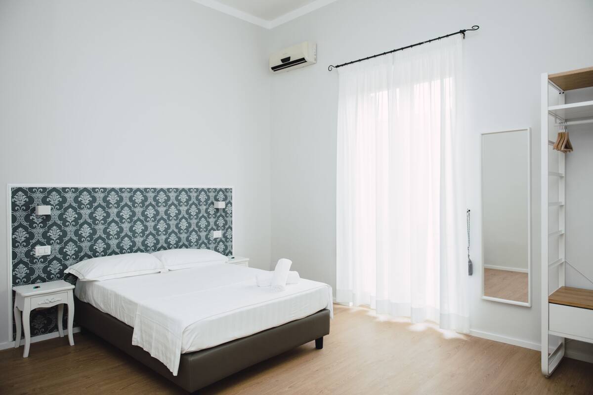 A modern bedroom with a bed, a nightstand, a wardrobe, and a window with sheer curtains.