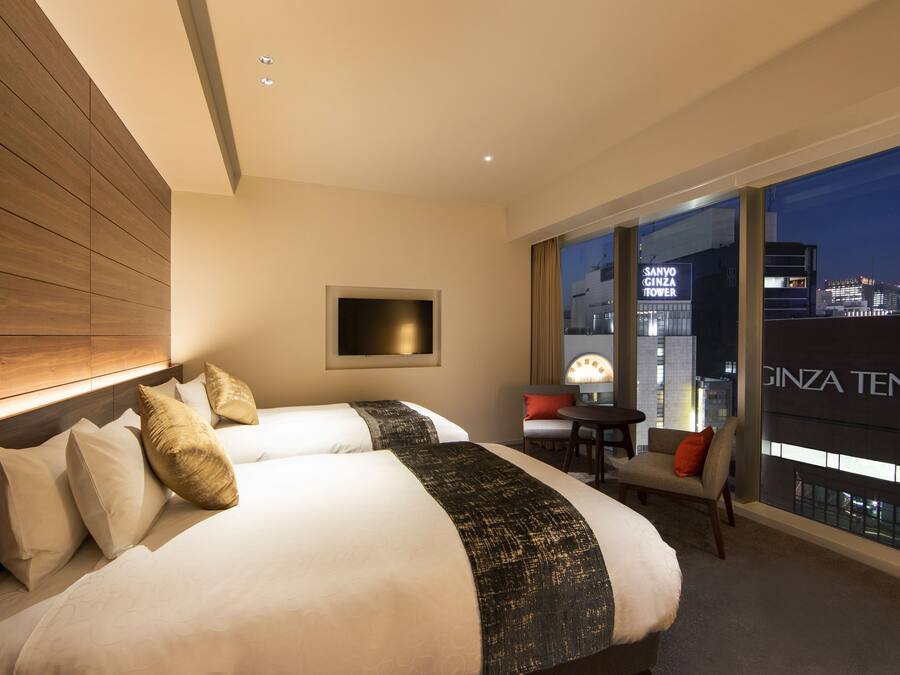 A hotel room with two beds, a TV, a chair, and a view of the city skyline.