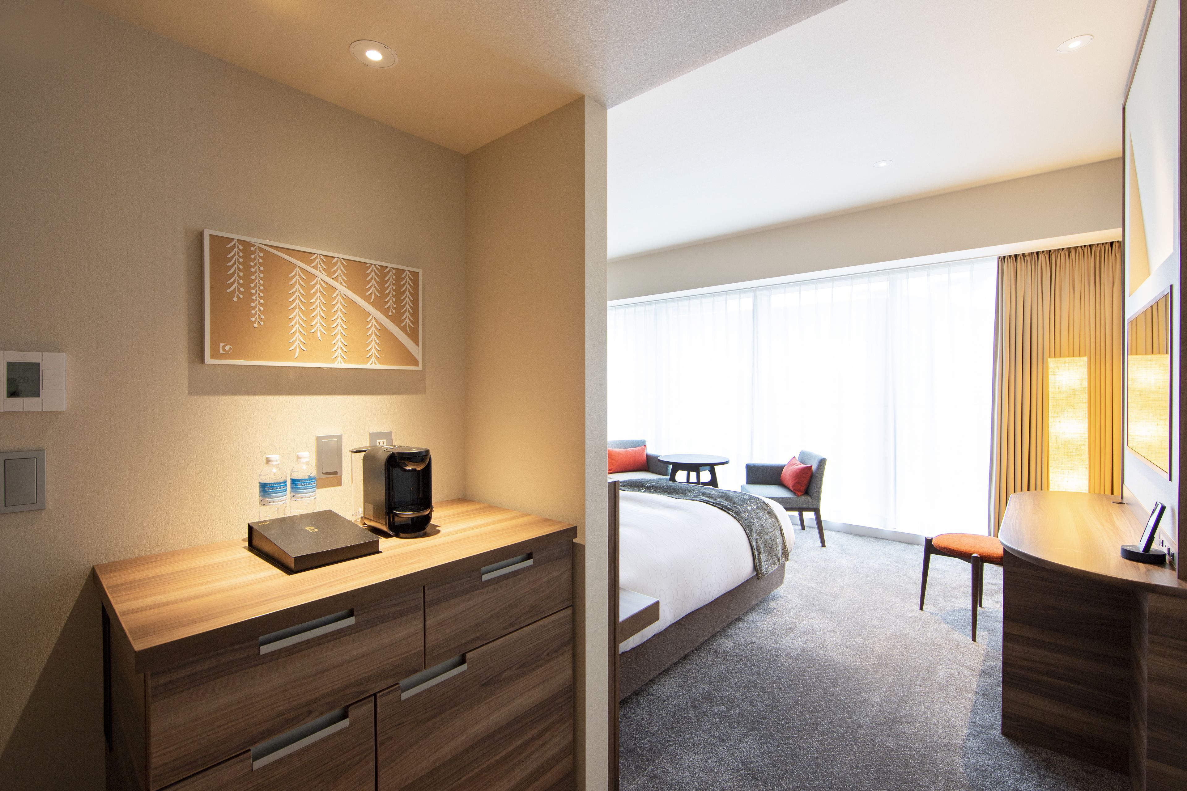 A hotel room with a wooden desk, a bed, a seating area with a table and chairs, and a large window with curtains.