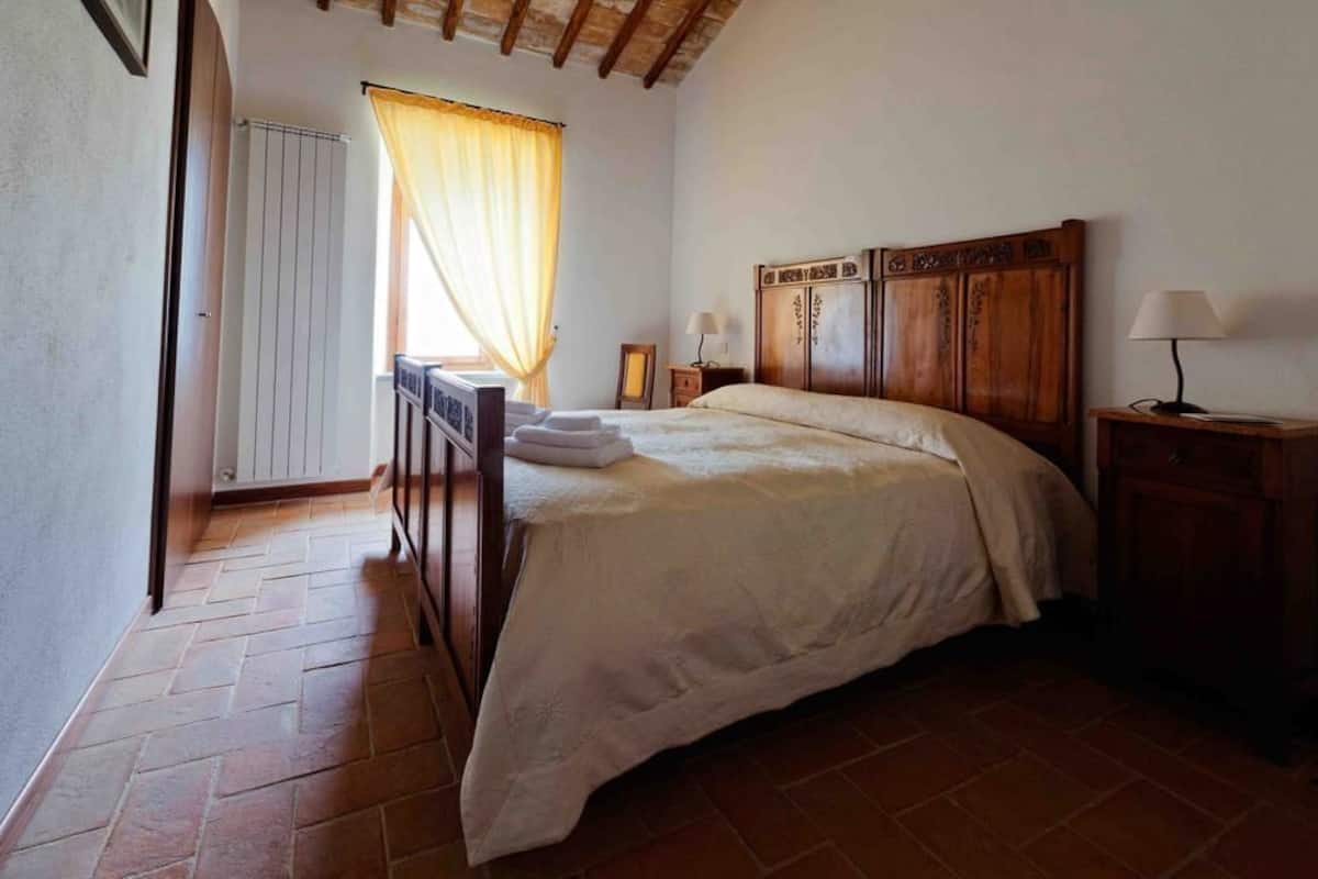 Un dormitorio con una cama de madera, un radiador, una ventana con cortinas y una mesita de noche con lámpara.