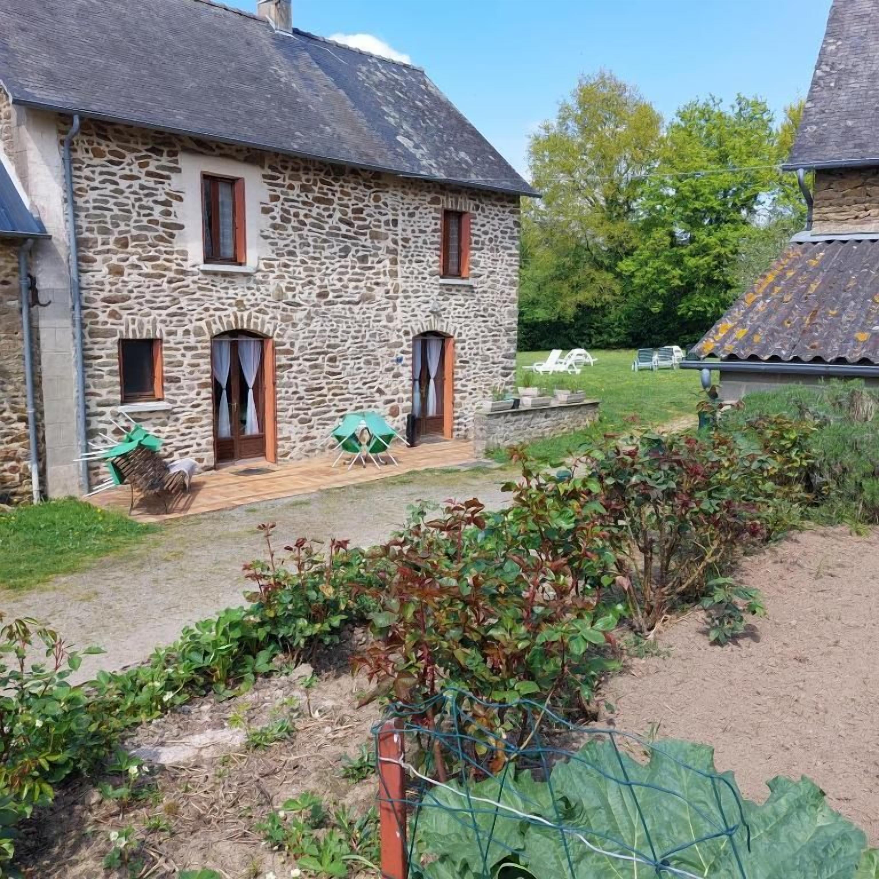 Een stenen huis met een houten terras, een tuin vol planten en een heldere lucht.