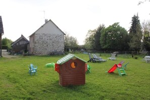 A grassy outdoor area with a wooden playhouse, a stone building, and outdoor furniture. - Maison d'hôtes La Lavandière (La Chapelle-au-Riboul)