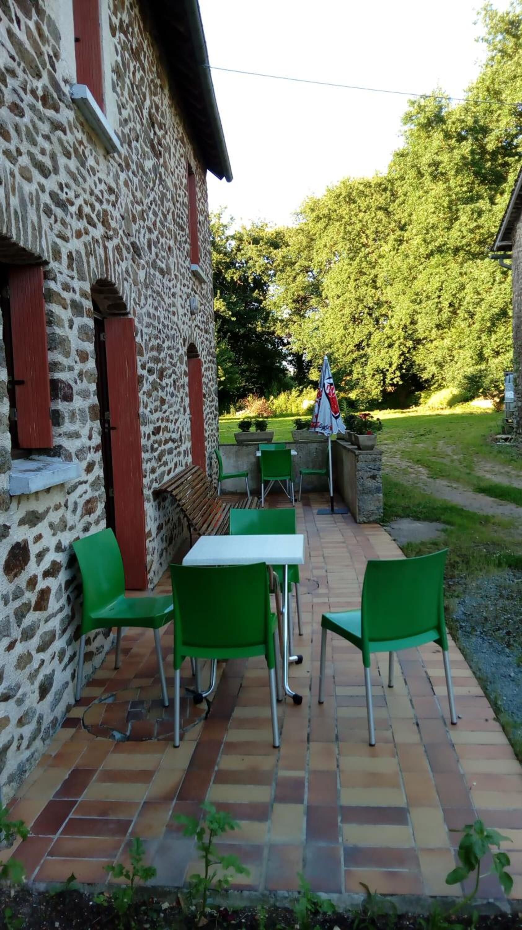 Un patio con sillas verdes y una mesa, un edificio de piedra con contraventanas rojas y un jardín con árboles y un sendero.