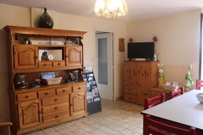 A dining area with a table and chairs, a television on a cabinet, and a wooden cabinet with drawers and shelves. - Maison d'hôtes La Lavandière (La Chapelle-au-Riboul)