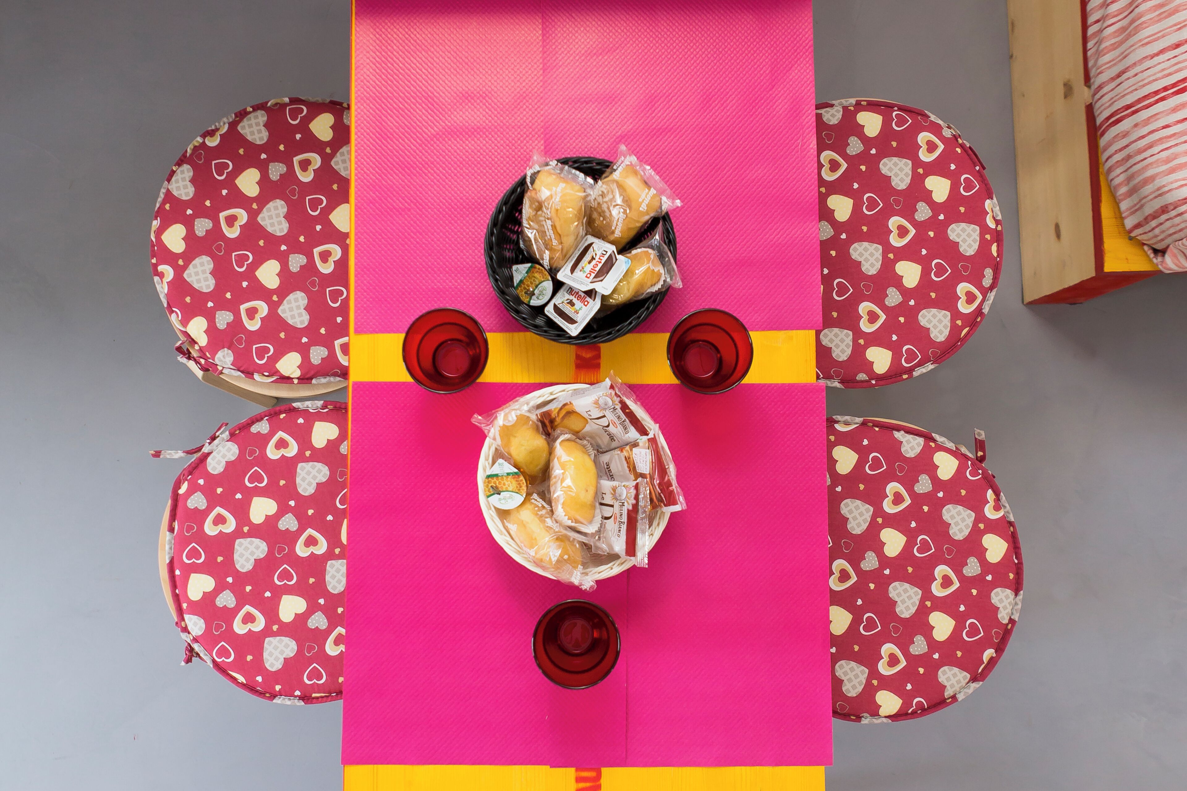 A table with heart-patterned cushions, a basket of pastries, and two glasses of red beverage.