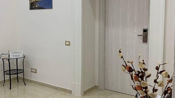 A hallway with a white door, a small table with a box labeled "THE", and a vase with artificial flowers. - B&B Napul'è (Naples)