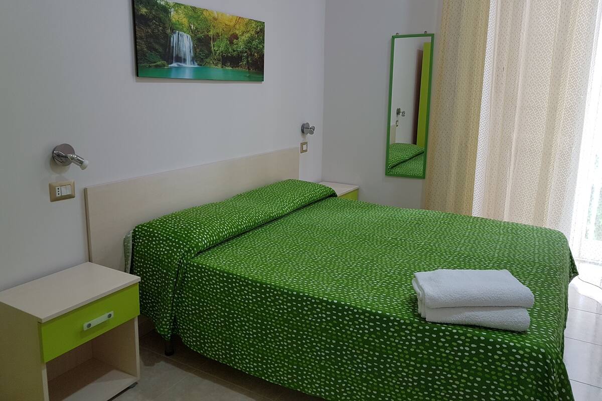 A neatly arranged bedroom with a green polka dot bedspread, a small bedside table, a wall-mounted mirror, and a painting of a waterfall.