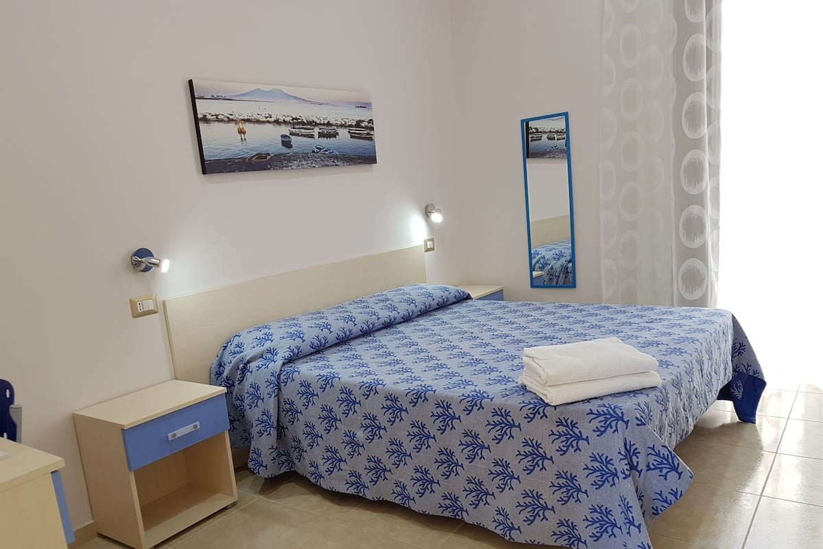 A bedroom with a bed, a nightstand, a drawer unit, a wall-mounted mirror, and a framed picture.