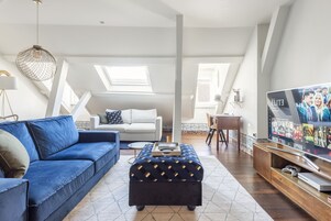 Uma sala de estar moderna com um sofá azul, um banquinho a combinar, uma televisão e um espaço de jantar.