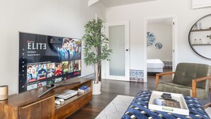 Un salon moderne avec un téléviseur à écran plat diffusant une série Netflix, un meuble de rangement en bois, une plante en pot, un fauteuil vert et un lit recouvert d’une couvre-lit à motifs.