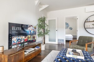 Uma sala de estar moderna com uma televisão de tela plana exibindo uma série da Netflix, um móvel para entretenimento de madeira, uma planta em vaso, uma poltrona verde e uma cama com colcha estampada.