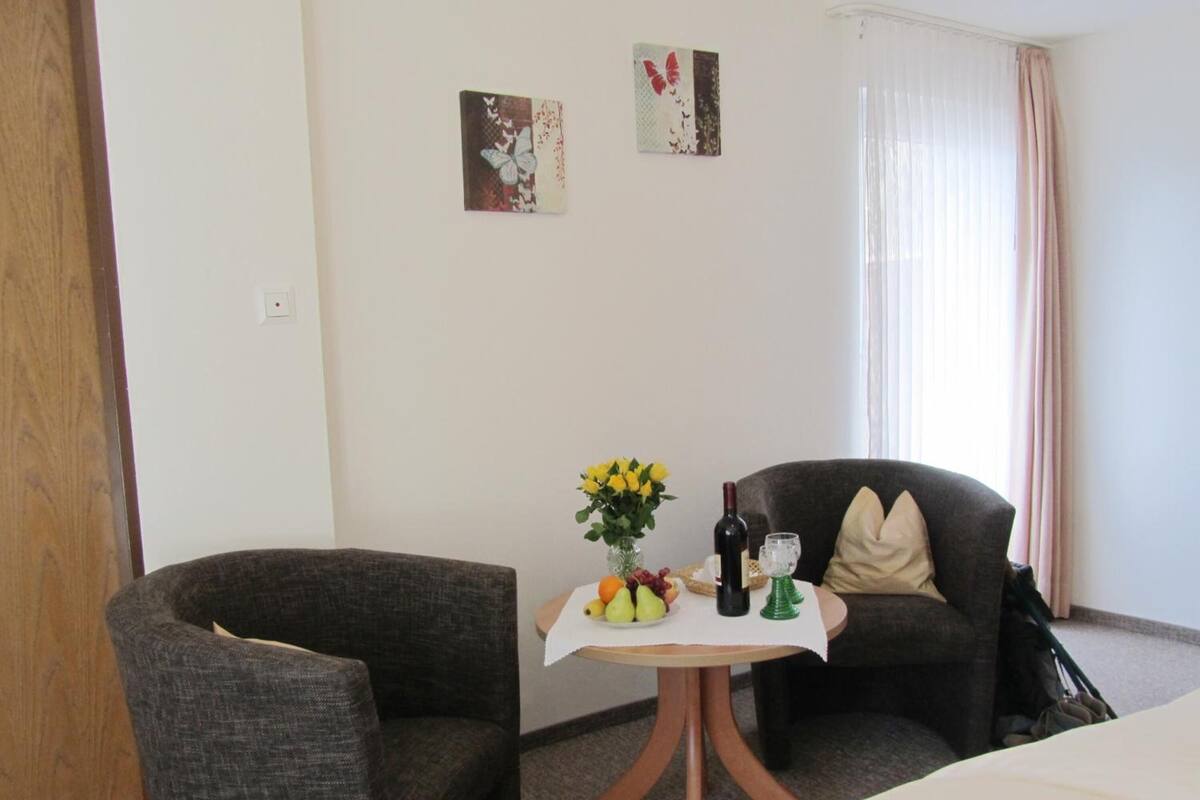 A hotel room with two grey armchairs, a small round table with a vase of yellow flowers, a bottle of wine, and a glass. There are two wall art pieces above the table.