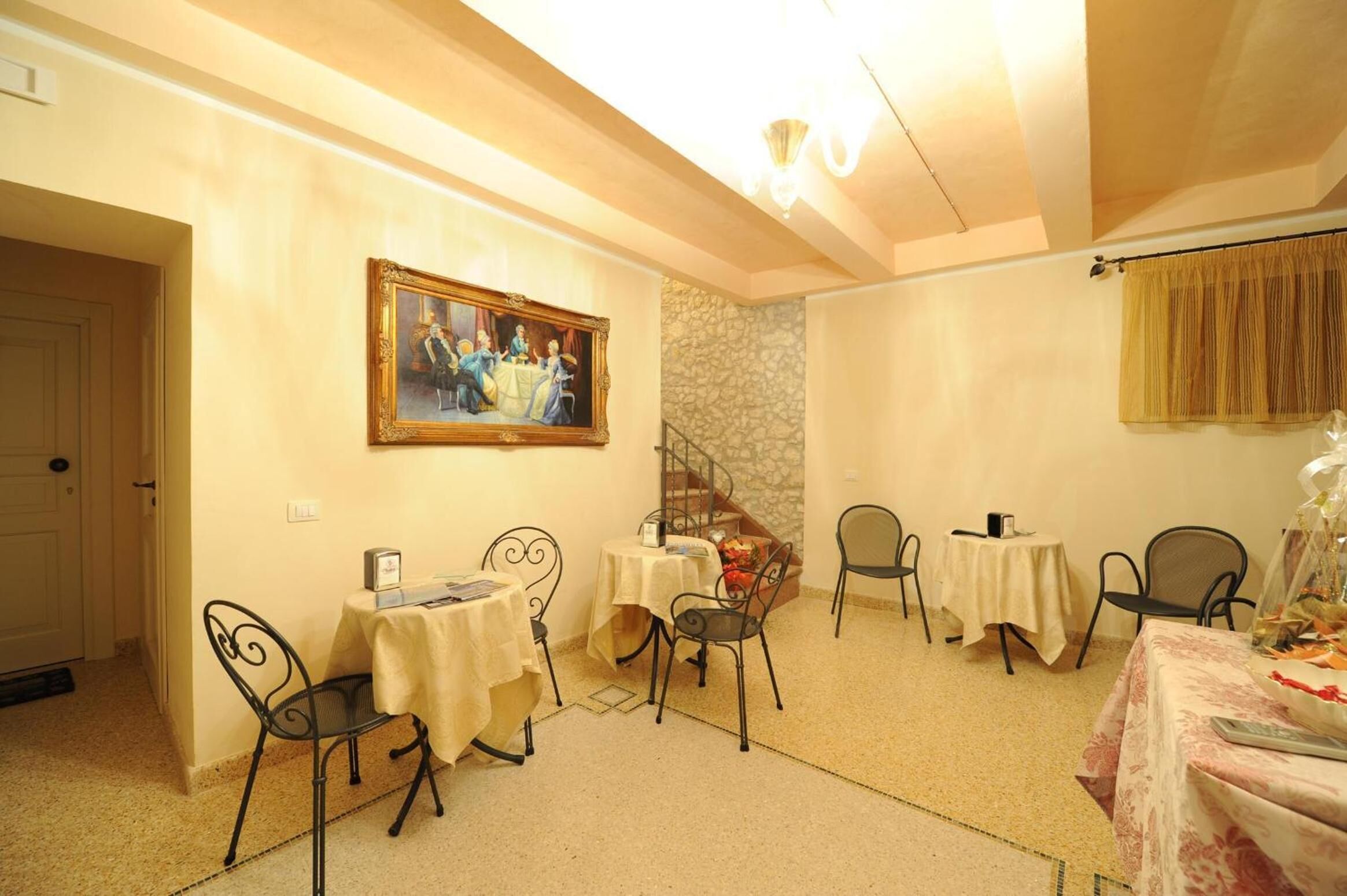 A dining area with four tables, each with a tablecloth and metal chairs. There is a staircase with a stone wall, a door, and a painting on the wall.