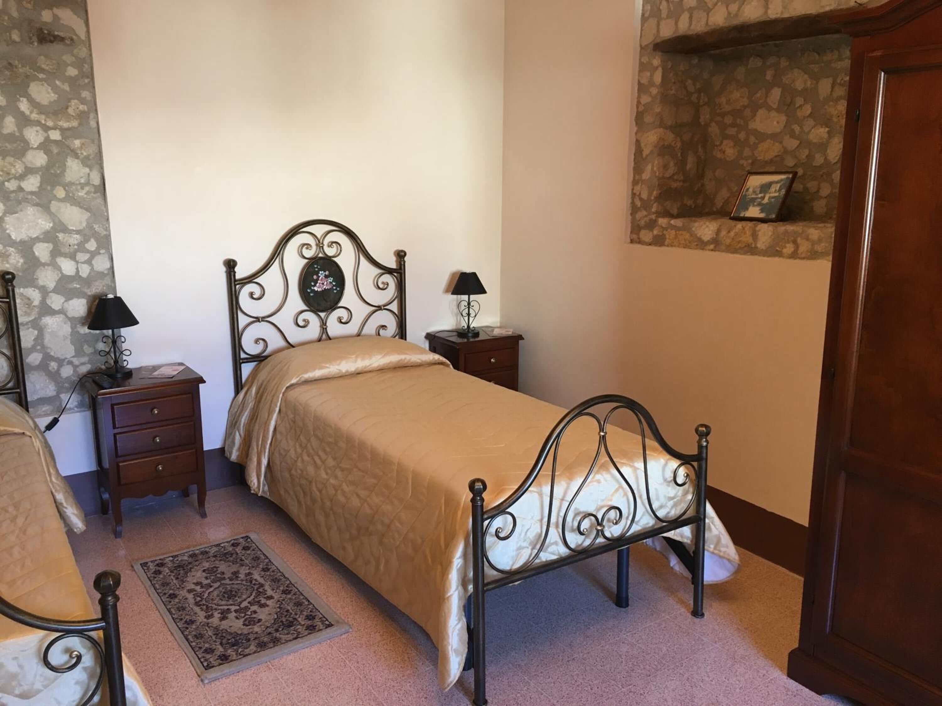 Un dormitorio con una cama, dos mesitas de noche, una butaca, una alfombra y una pared de piedra.