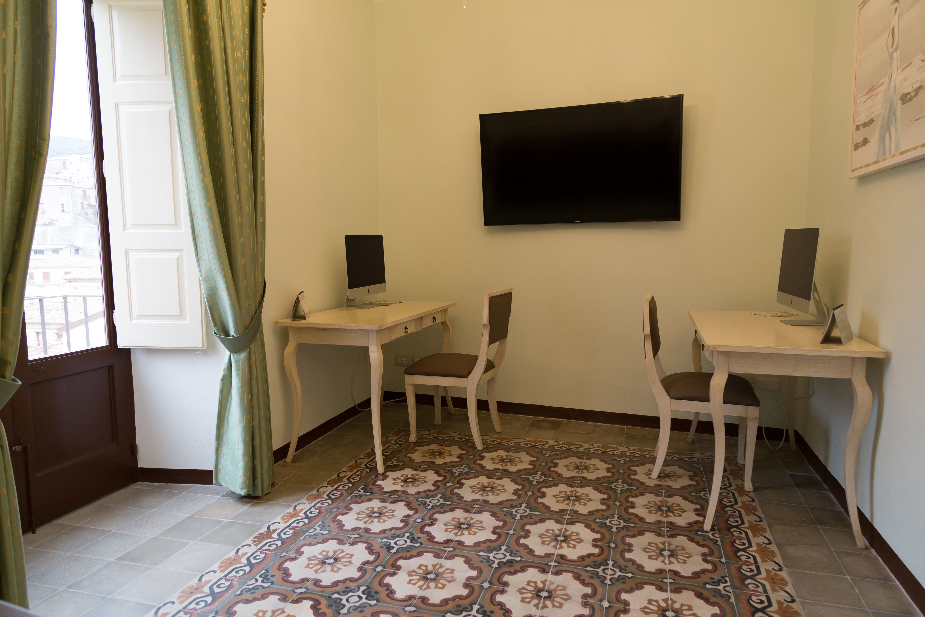 Ein Zimmer mit zwei Schreibtischen, jeweils mit einem Computer und einem Stuhl, einem Flachbildfernseher an der Wand, gemusterten Bodenfliesen und einem Fenster mit Vorhängen.
