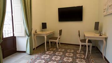 Ein Zimmer mit zwei Schreibtischen, jeweils mit einem Computer und einem Stuhl, einem Flachbildfernseher an der Wand, gemusterten Bodenfliesen und einem Fenster mit Vorhängen.
