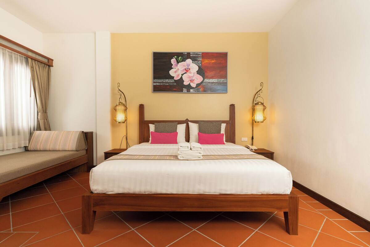 A neatly made bed with a wooden frame, a headboard with a decorative light fixture, and a painting of flowers on the wall.