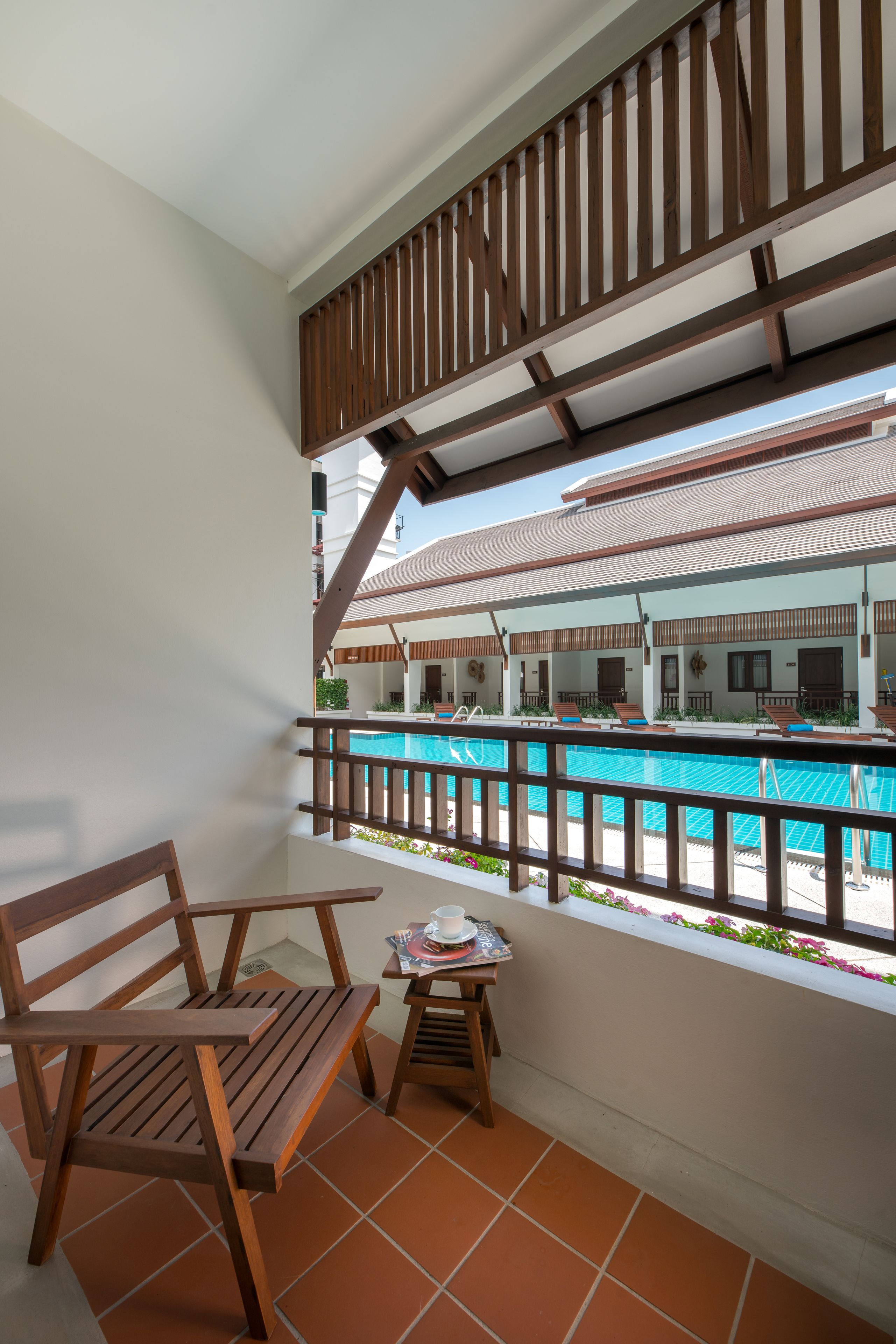 A balcony with a wooden chair, a small table with a cup and saucer, and a view of a pool area with lounge chairs and umbrellas.