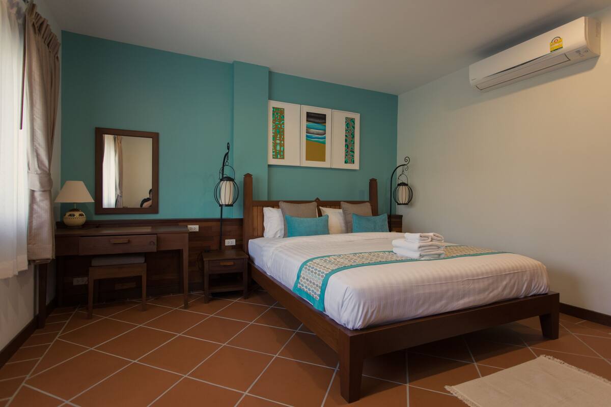 A bedroom with a bed, a desk, a chair, a nightstand, a mirror, and a wall-mounted air conditioner.