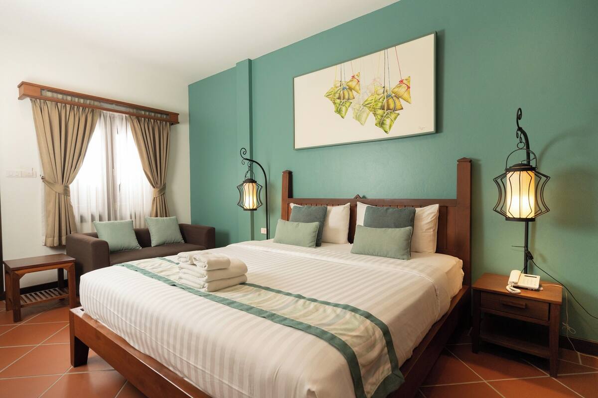 A hotel room with a large bed, a sofa, a nightstand with a lamp, and a painting on the wall.