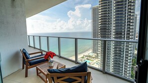 A balcony with two wooden chairs, a small table, and a red flower vase. A large window offers a view of the ocean and a high-rise building. - Lyfe Residence Building - Private apts over the beach (Hollywood)