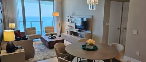 Un salon moderne avec une table à manger ronde, des chaises de couleur beige, un lustre, un téléviseur à écran plat et une grande fenêtre donnant sur la mer.
