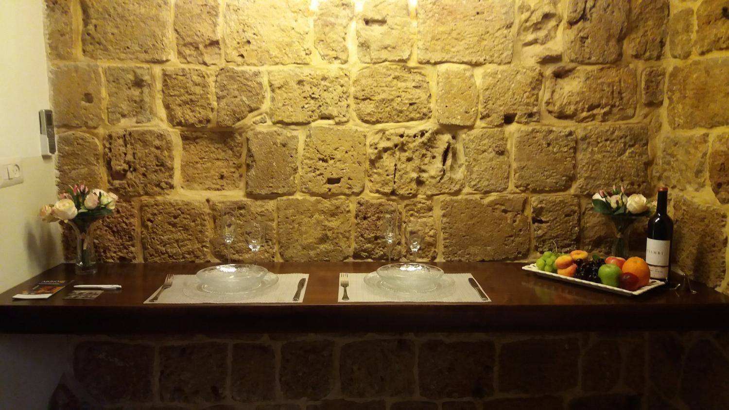 Pared de piedra con una barra de madera que presenta dos cuencos de vidrio transparente, un plato de fruta y una botella de vino.