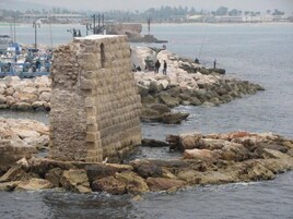 Una construcción de piedra cerca de una costa rocosa, con barcos al fondo.