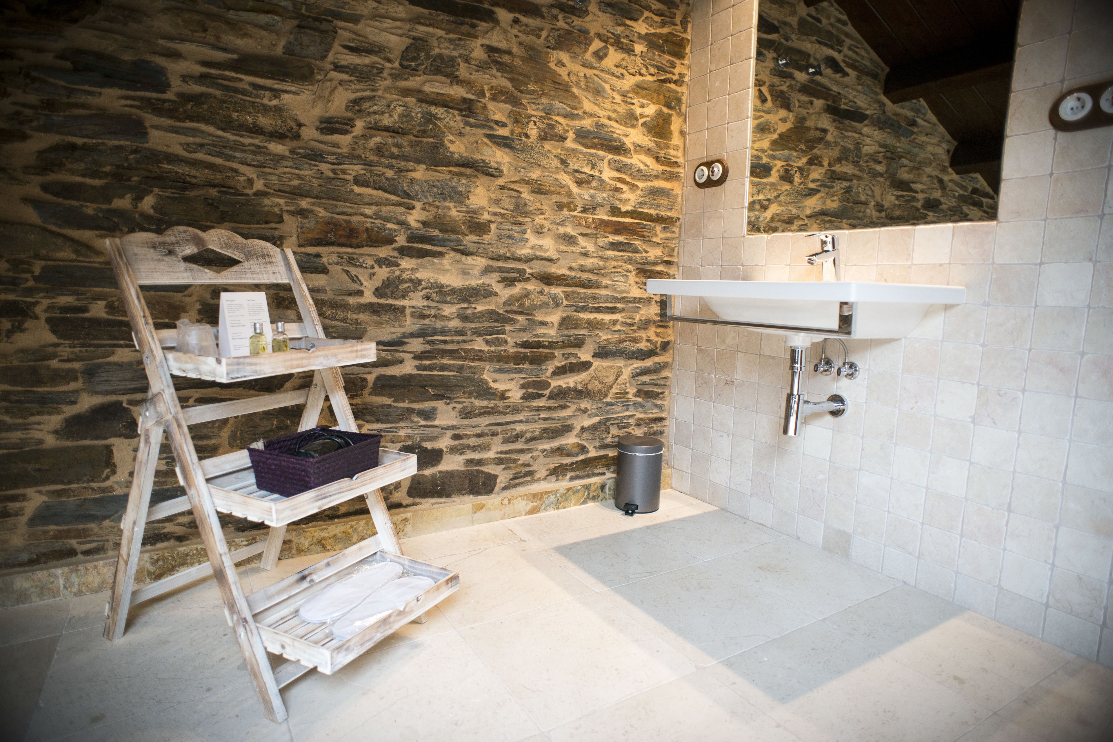 A bathroom with a stone wall, a wooden shelf with toiletries, a sink, and a trash can.