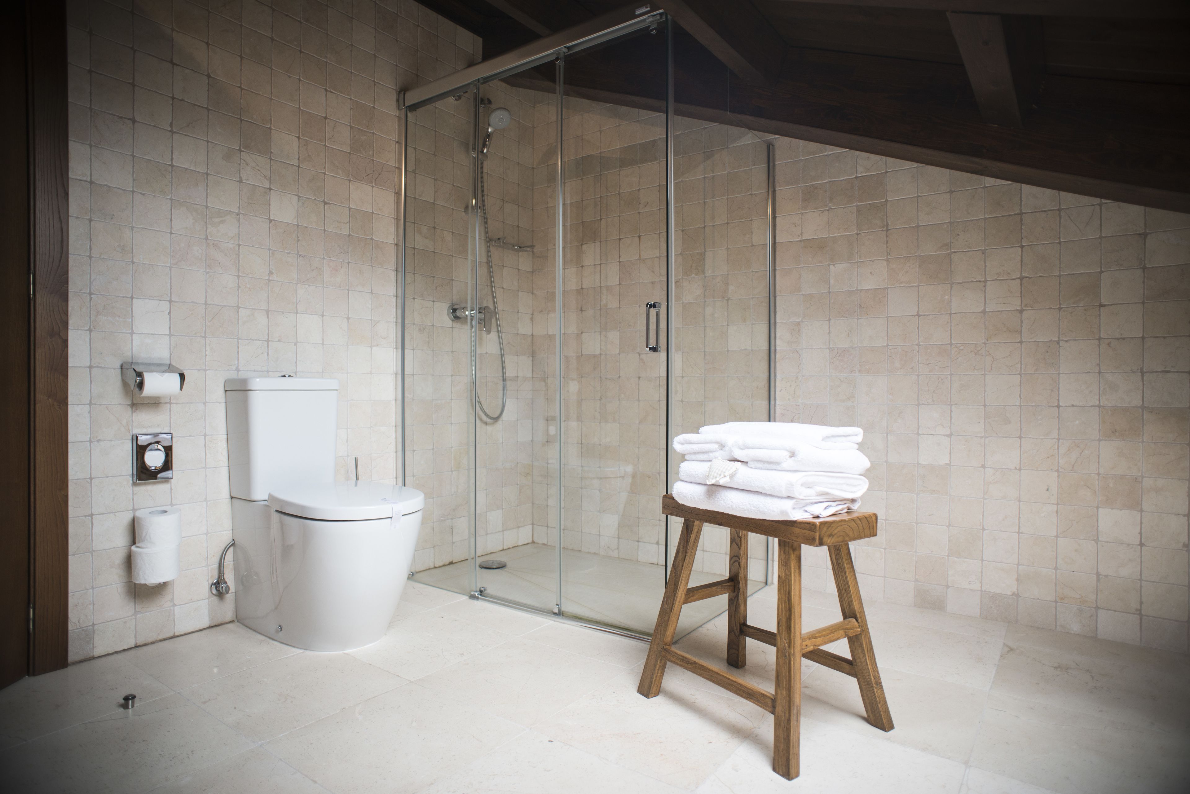 Un baño con cabina de ducha de vidrio, un inodoro y un taburete de madera con toallas dobladas.
