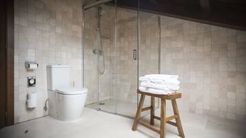 Un baño con cabina de ducha de vidrio, un inodoro y un taburete de madera con toallas dobladas.