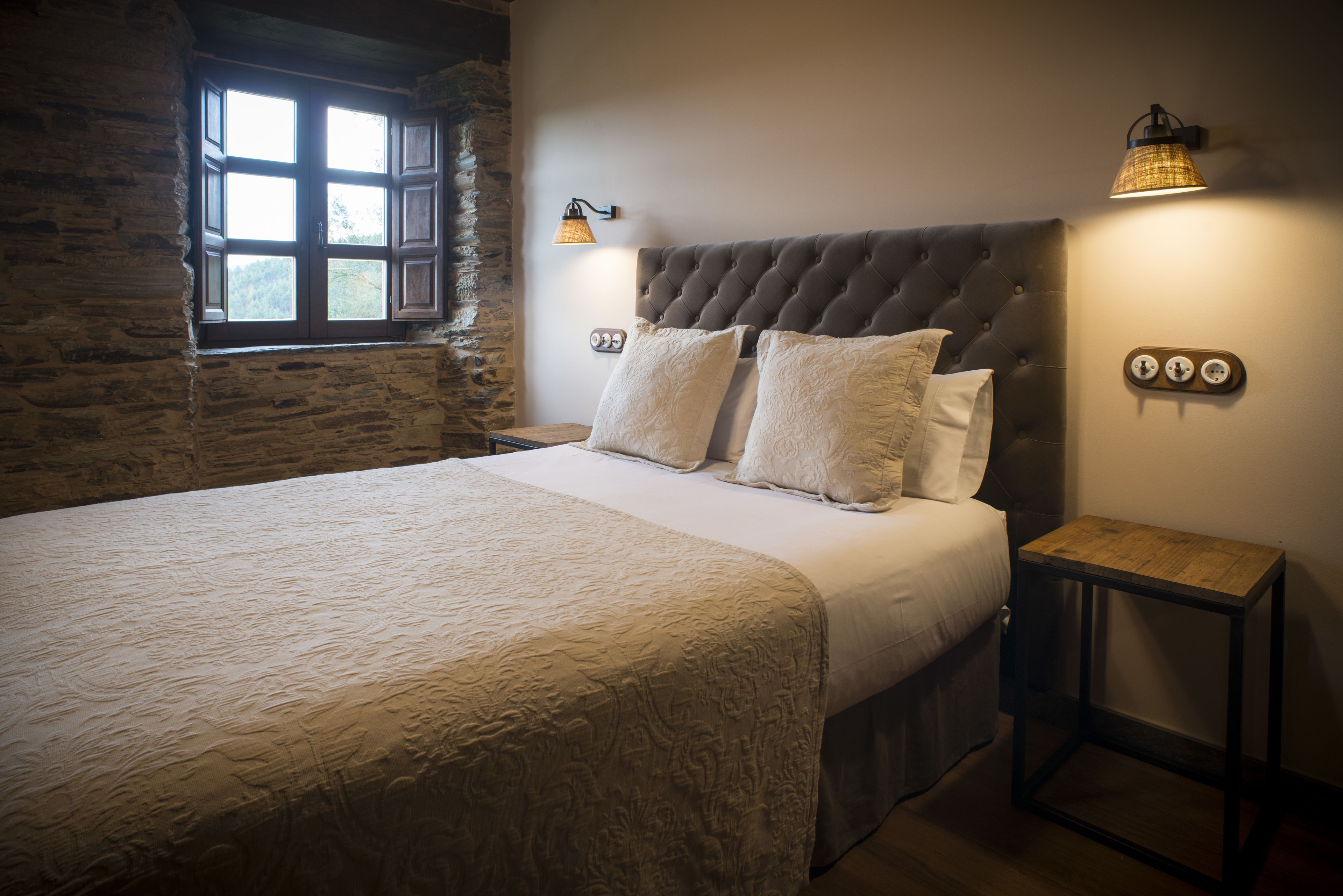 A bedroom with a large bed, a stone wall, a window, and a nightstand with a lamp.