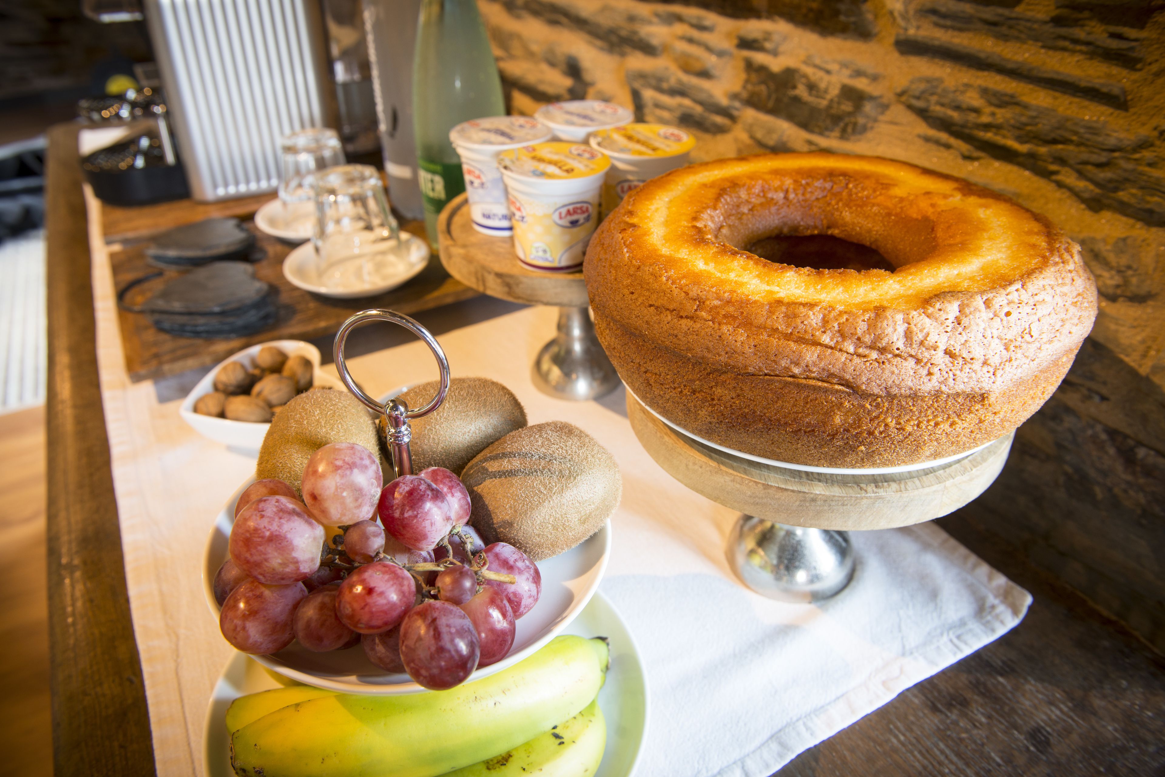 Uma mesa com um bolo bundt, um prato de kiwis, uvas e bananas, e uma bandeja com copos de iogurte.