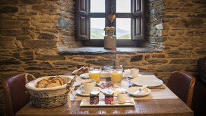 Mesa de desayuno dispuesta con pan, huevos, tocino y bebidas en una habitación de paredes de piedra con vistas a las montañas.