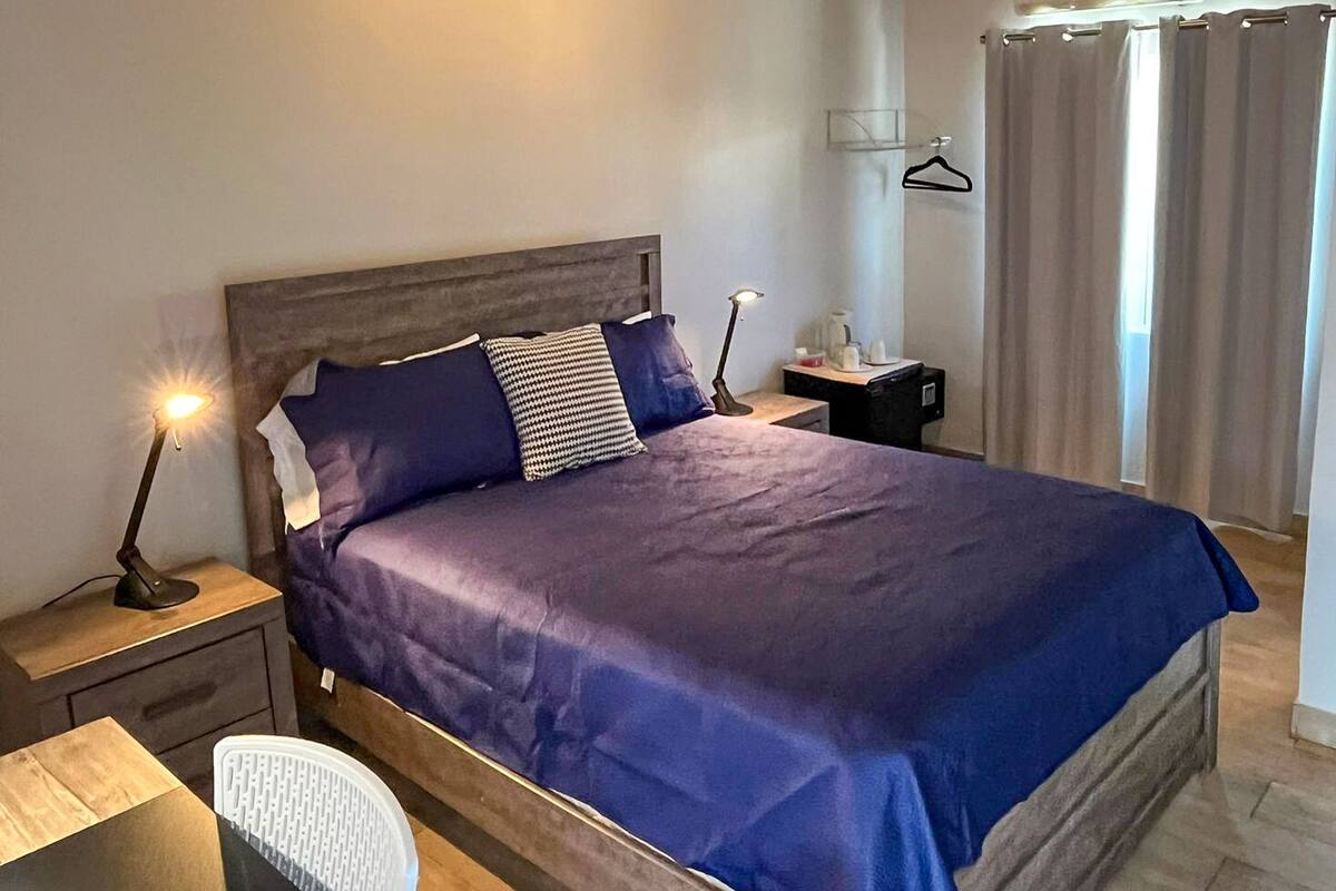 A neatly made bed with a blue bedspread and pillows, a bedside table with a lamp, a chair, and a small nightstand with a bottle and a cup.