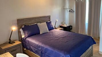 A neatly made bed with a blue bedspread and pillows, a bedside table with a lamp, a chair, and a small nightstand with a bottle and a cup.