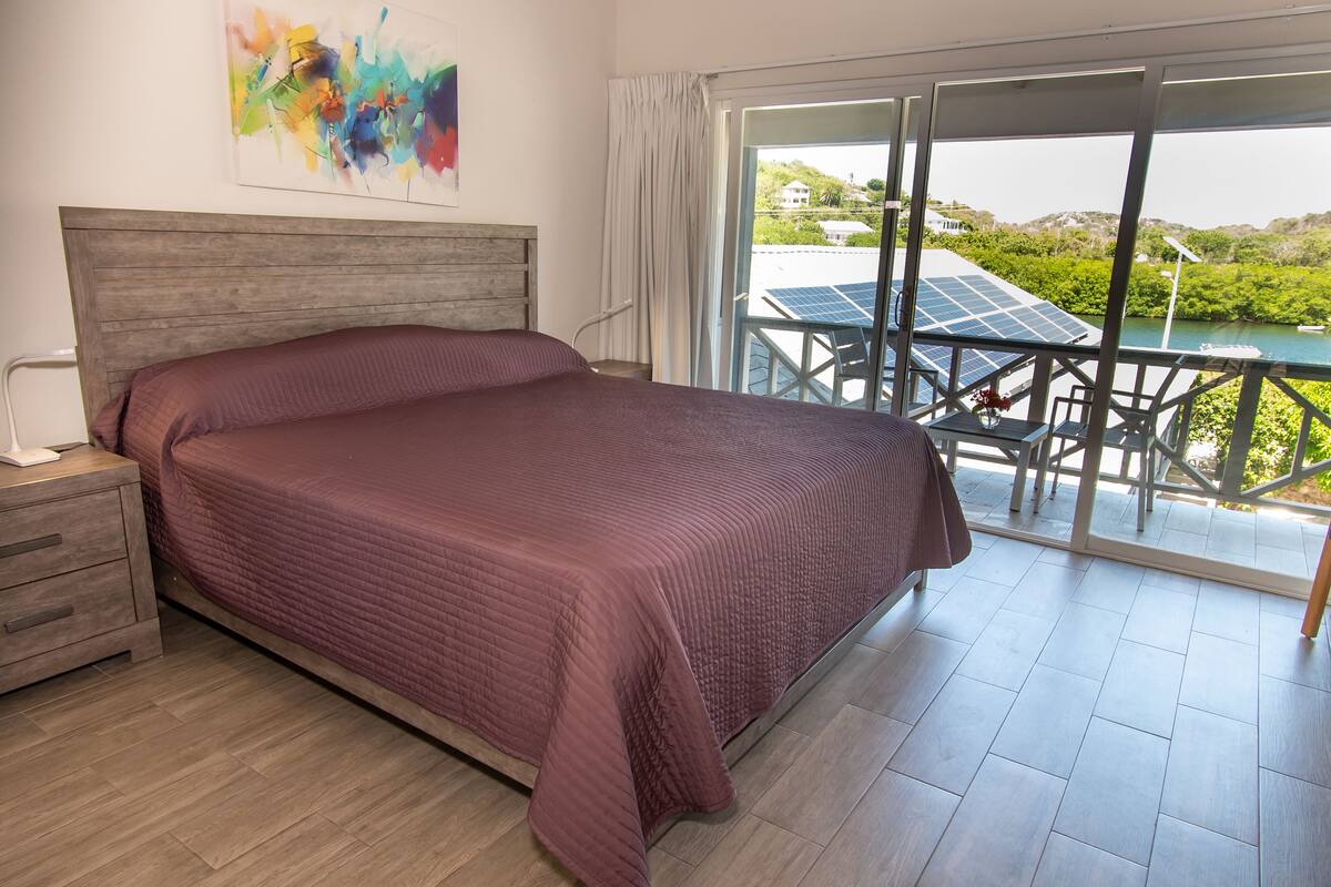 A bedroom with a large bed, a nightstand, a painting on the wall, and a view of a balcony with solar panels and a boat.