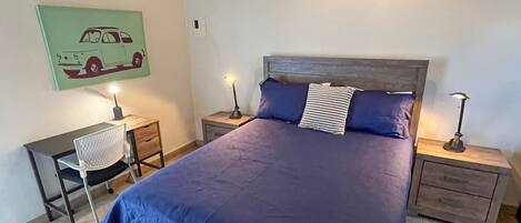 Un dormitorio con una cama grande, dos mesitas de noche con lámparas, una silla y una obra de arte colgada en la pared.