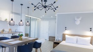A modern hotel room with a wooden bed, a dining area with a table and chairs, a kitchen with white cabinets, and a ceiling light fixture. - Royal Suite near Lake (Sumy)