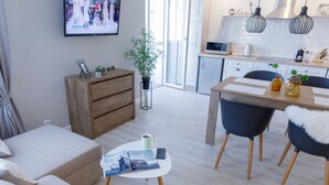 A modern living room with a sofa, a coffee table with magazines, a TV displaying a fashion show, a wooden cabinet, and a dining area with chairs and a table. - Royal Suite near Lake (Sumy)
