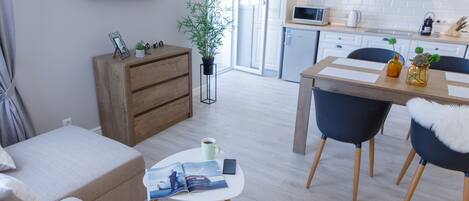 A modern living room with a sofa, a coffee table with magazines, a TV displaying a fashion show, a wooden cabinet, and a dining area with chairs and a table.
