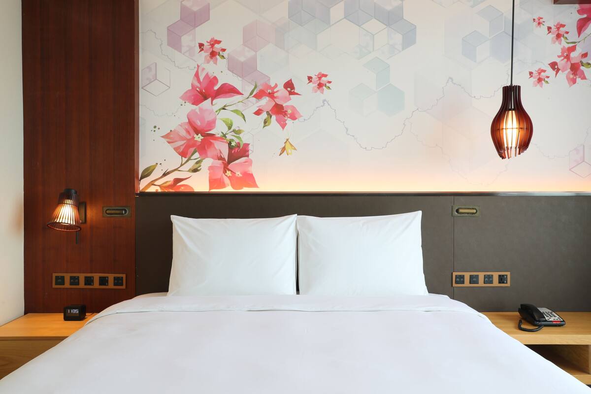 Ein Hotelzimmer mit Bett, Nachttisch mit Telefon, Wanduhr und dekorativer Wand mit geometrischen Mustern und Blumen.