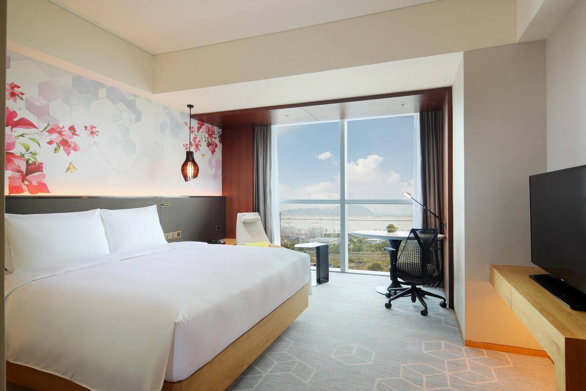 Ein Hotelzimmer mit einem großen Bett, einem Schreibtisch mit Fernseher, einem Sessel und Blick auf eine Stadtkulisse durch große Fenster.