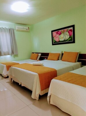 A hotel room with three single beds, each with a white and orange bedspread. There is a painting of flowers on the wall, and an air conditioning unit mounted on the wall.