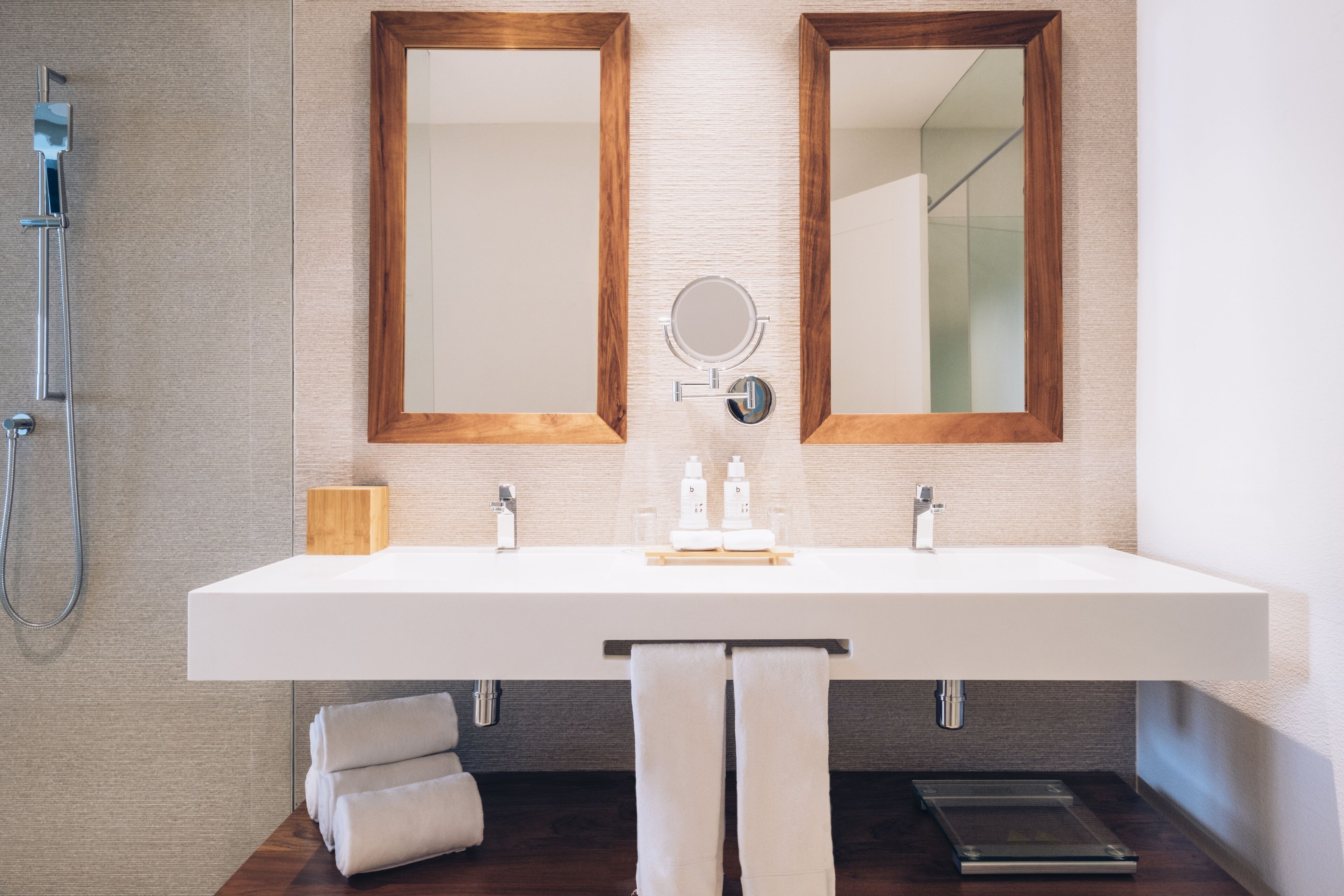 A modern bathroom with a large white sink, two wooden-framed mirrors, a handheld showerhead, and a small wooden box on the counter.