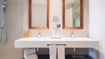 A modern bathroom with a large white sink, two wooden-framed mirrors, a handheld showerhead, and a small wooden box on the counter.