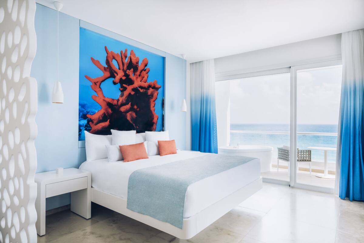 A modern hotel room with a large bed, a view of the ocean, and a tropical-themed wall mural.